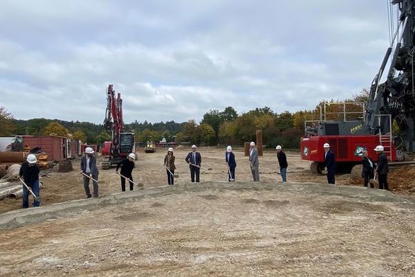 Zehn Personen mit Spaten und einem Bagger im Hintergrund, werfen den Sand